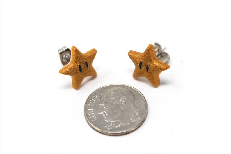May include: A pair of gold star-shaped earrings with black eyes. The earrings are made of polymer clay and have a shiny finish. The earrings are about the size of a dime.