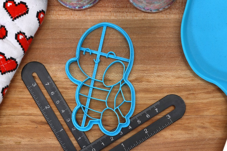 May include: A blue plastic cookie cutter shaped like a cartoon turtle with a smiling face. The cutter has a grid pattern on the shell.