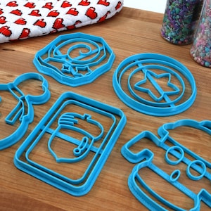 May include: A set of six blue plastic cookie cutters shaped like a snowman, a star, a present, a mitten, a wreath, and a shield with a star in the center.
