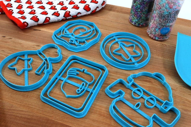 May include: A set of six blue plastic cookie cutters shaped like a snowman, a star, a present, a mitten, a wreath, and a shield with a star in the center.