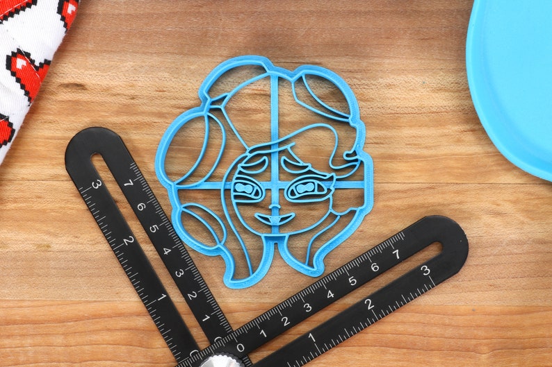 May include: A blue plastic cookie cutter shaped like a cartoon character with long hair and large eyes.