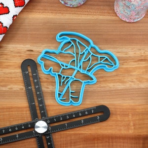 May include: A blue plastic cookie cutter shaped like a bunch of broccoli. The cutter is on a wooden surface with a ruler and other baking supplies.