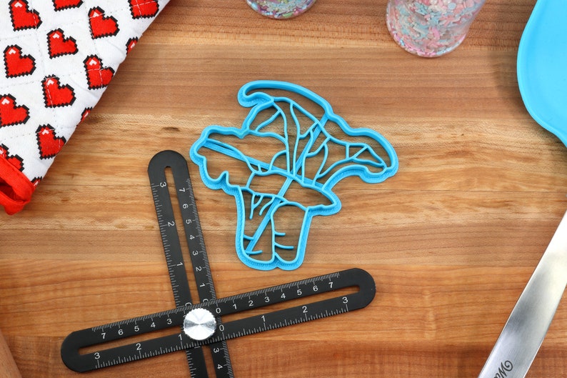 May include: A blue plastic cookie cutter shaped like a bunch of broccoli. The cutter is on a wooden surface with a ruler and other baking supplies.