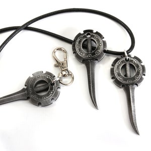 May include: Three silver-colored metal pendants with a circular design and a pointed tip. The pendants are attached to a black cord.