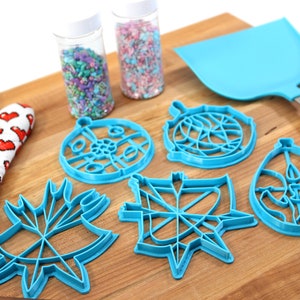 May include: A set of six blue plastic cookie cutters shaped like ornaments, a spatula, and two jars of sprinkles. The cookie cutters are on a wooden surface.