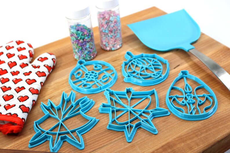 May include: A set of six blue plastic cookie cutters shaped like ornaments, a spatula, and two jars of sprinkles. The cookie cutters are on a wooden surface.