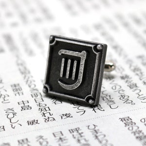 Puede incluir: Gemelos de plata con un dise&ntilde;o cuadrado negro que presenta un s&iacute;mbolo de lira plateado.