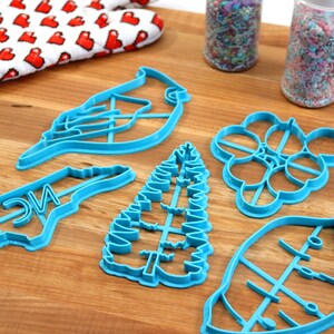May include: A set of six blue plastic cookie cutters on a wooden surface. The cutters are shaped like a cardinal, a tree, a leaf, a bunch of grapes, a state of North Carolina, and a generic leaf.