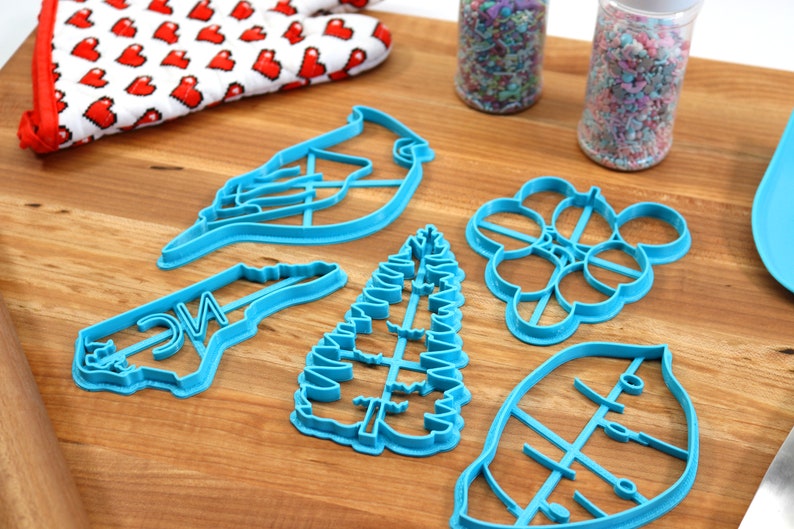 May include: A set of six blue plastic cookie cutters on a wooden surface. The cutters are shaped like a cardinal, a tree, a leaf, a bunch of grapes, a state of North Carolina, and a generic leaf.
