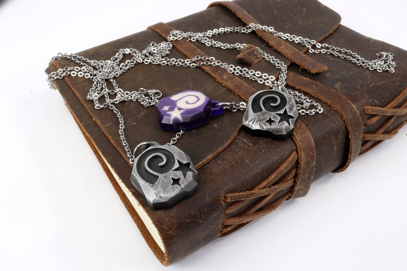 May include: Three silver necklaces with pendants featuring a spiral design and stars. The pendants are on a brown leather journal.