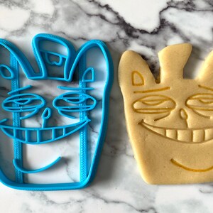 May include: A blue plastic cookie cutter and a baked cookie, both shaped like a cartoon face with a wide smile and closed eyes.