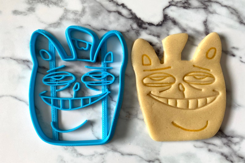 May include: A blue plastic cookie cutter and a baked cookie, both shaped like a cartoon face with a wide smile and closed eyes.