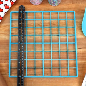 May include: A blue plastic grid with 36 squares, a black ruler, a wooden rolling pin, and a blue spatula with a white handle.