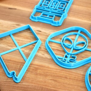 May include: A set of five blue plastic cookie cutters. The cutters are shaped like a kite, an alien head, a building, a cloud with the word "Dream" on it, and a house.