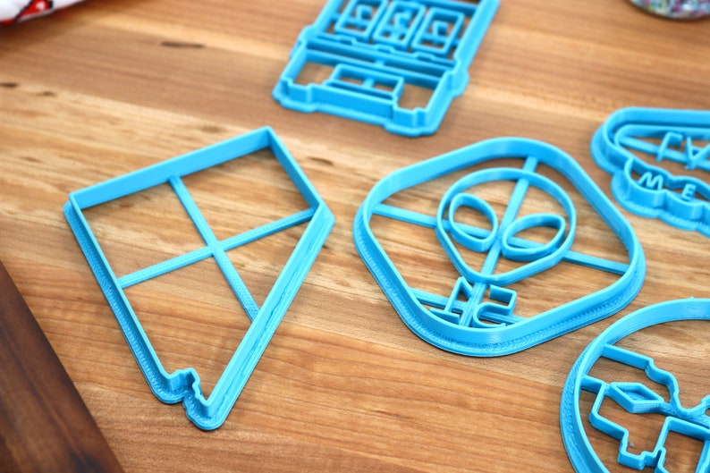 May include: A set of five blue plastic cookie cutters. The cutters are shaped like a kite, an alien head, a building, a cloud with the word "Dream" on it, and a house.