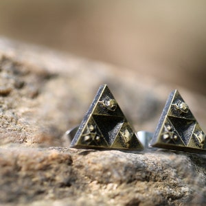 May include: A pair of gold and black triangle-shaped earrings with a detailed design. The earrings are on a brown, textured surface.