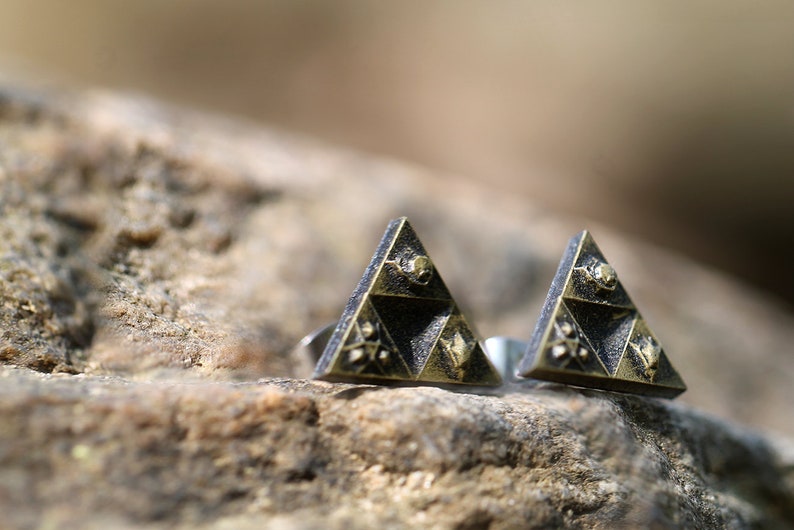 May include: A pair of gold and black triangle-shaped earrings with a detailed design. The earrings are on a brown, textured surface.