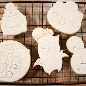 May include: Five unbaked sugar cookies on a wire rack. The cookies are shaped like cartoon characters with different expressions.