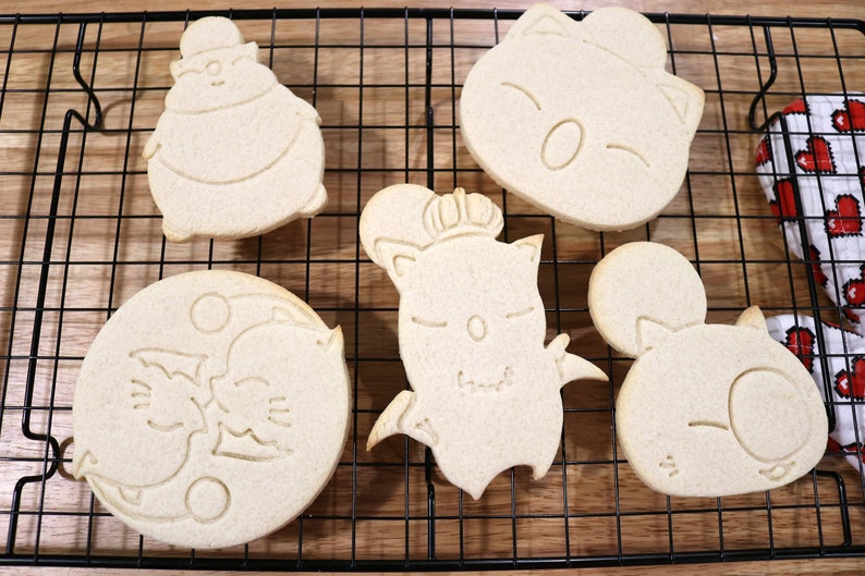 May include: Five unbaked sugar cookies on a wire rack. The cookies are shaped like cartoon characters with different expressions.