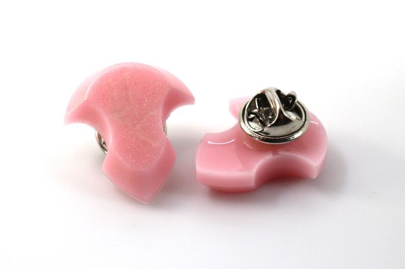 Puede incluir: Dos pines de resina rosa con forma de media luna estilizada. El pin de la derecha tiene un cierre de pin de metal plateado.