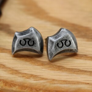May include: A pair of silver earrings with a geometric design and a black symbol. The earrings are on a wooden surface.