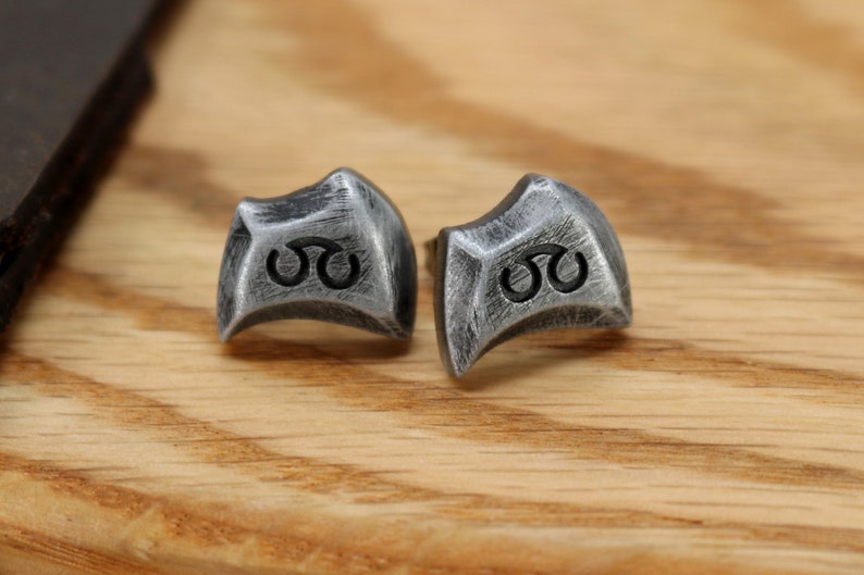 May include: A pair of silver earrings with a geometric design and a black symbol. The earrings are on a wooden surface.