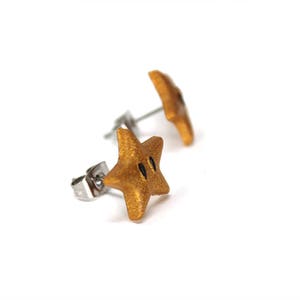 May include: A pair of gold star-shaped earrings with black accents. The earrings are made of polymer clay and have a shiny finish.