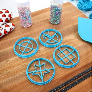 May include: A collection of baking tools and supplies on a wooden surface. Includes blue cookie cutters in various geometric shapes, a blue spatula, a Wilton ruler, a heart-patterned oven mitt, and jars of sprinkles.