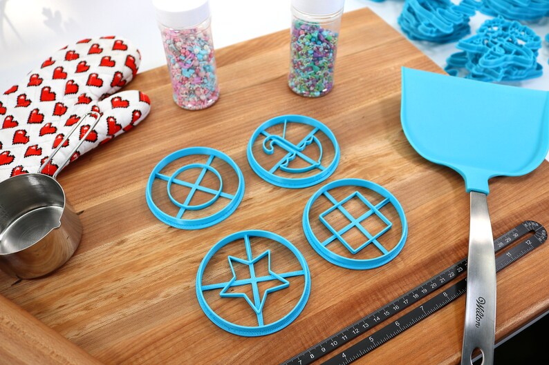 May include: A collection of baking tools and supplies on a wooden surface. Includes blue cookie cutters in various geometric shapes, a blue spatula, a Wilton ruler, a heart-patterned oven mitt, and jars of sprinkles.