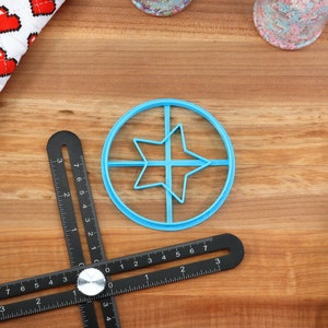 May include: A light blue star-shaped cookie cutter inside a circle, a black multi-angle measuring tool, and a white and red heart-patterned kitchen towel are on a wooden surface. Sprinkles and a blue spatula are also visible.