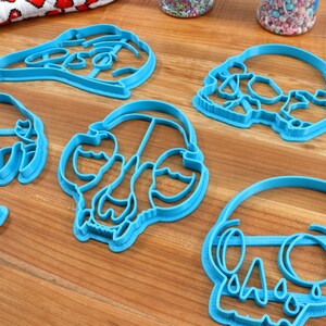 May include: Five blue plastic cookie cutters shaped like skulls. The cutters are on a wooden surface.