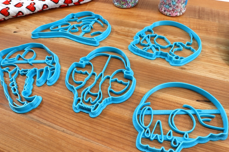 May include: Five blue plastic cookie cutters shaped like skulls. The cutters are on a wooden surface.