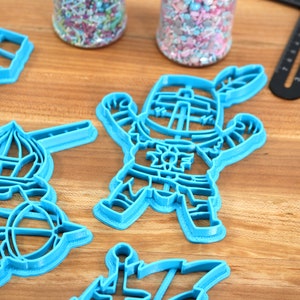 May include: Four blue plastic cookie cutters on a wooden surface. The cutters are shaped like a knight, a hot air balloon, a castle, and a ship.
