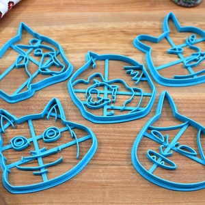 May include: Five blue plastic cookie cutters shaped like cat faces. The cutters have a grid pattern inside the outline of the cat faces.
