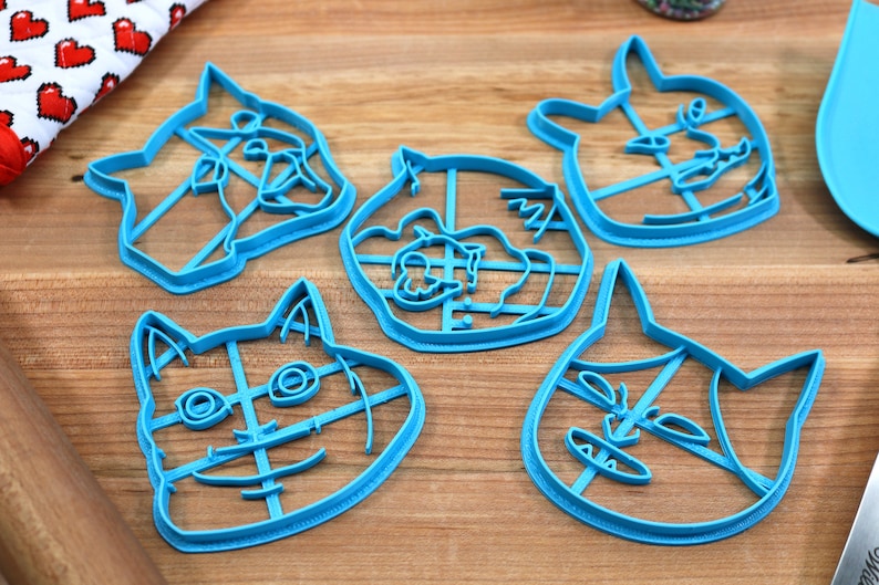 May include: Five blue plastic cookie cutters shaped like cat faces. The cutters have a grid pattern inside the outline of the cat faces.