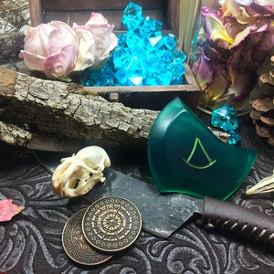 May include: A green resin pendant with a triangular symbol, two antique-looking coins, a small animal skull, and a knife with a leather handle. The pendant is surrounded by dried flowers and a pine cone.