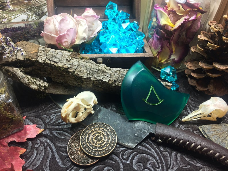 May include: A green resin pendant with a triangular symbol, two antique-looking coins, a small animal skull, and a knife with a leather handle. The pendant is surrounded by dried flowers and a pine cone.