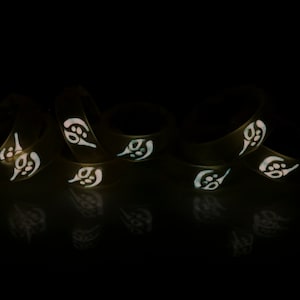 May include: A group of six black silicone rings with a white glow-in-the-dark design. The rings are stacked on top of each other and are illuminated in the dark.