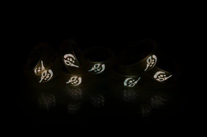May include: A group of six black silicone rings with a white glow-in-the-dark design. The rings are stacked on top of each other and are illuminated in the dark.