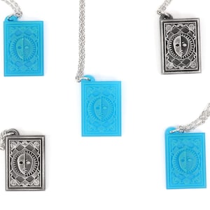 Puede incluir: Cuatro collares con colgantes que presentan un dise&ntilde;o plateado y azul de una cara con un sol y una luna a cada lado. Los colgantes son rectangulares y tienen un borde detallado.