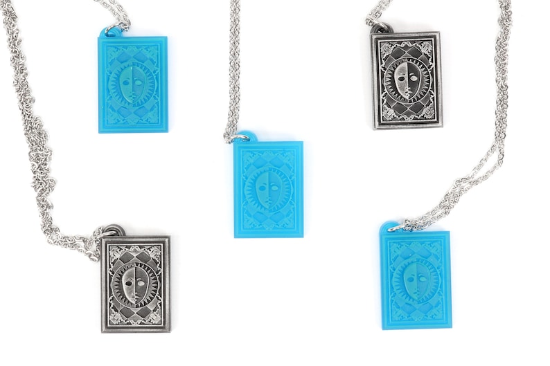 Puede incluir: Cuatro collares con colgantes que presentan un dise&ntilde;o plateado y azul de una cara con un sol y una luna a cada lado. Los colgantes son rectangulares y tienen un borde detallado.