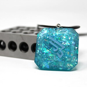 May include: A turquoise blue pendant necklace with a silver chain and a clear resin pendant with a white geometric design and iridescent glitter.