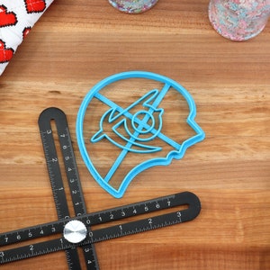 May include: A blue plastic cookie cutter shaped like a human head with a stylized design inside. The cutter is on a wooden surface.