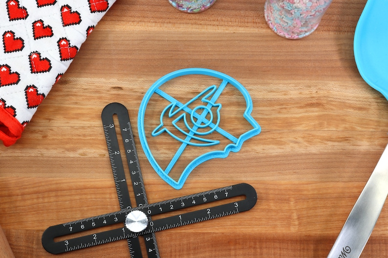 May include: A blue plastic cookie cutter shaped like a human head with a stylized design inside. The cutter is on a wooden surface.