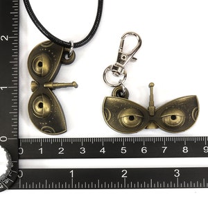May include: Two gold-colored robot eye charms, one on a black cord necklace and the other on a silver key chain. Both charms feature large, round eyes with black pupils and a small antenna on top.