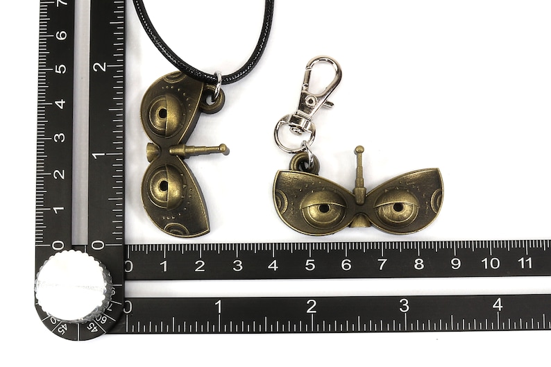 May include: Two gold-colored robot eye charms, one on a black cord necklace and the other on a silver key chain. Both charms feature large, round eyes with black pupils and a small antenna on top.