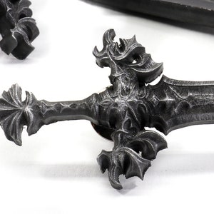May include: A detailed, ornate, silver-colored sword pin with a pointed tip and intricate designs. The pin is made of metal and has a black finish.