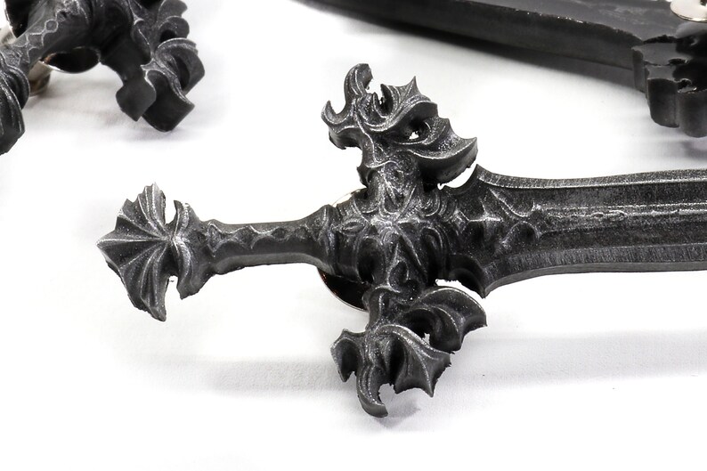 May include: A detailed, ornate, silver-colored sword pin with a pointed tip and intricate designs. The pin is made of metal and has a black finish.