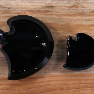 May include: Two black resin crescent moon shapes, one larger than the other. The larger crescent moon has gold flecks embedded in the resin.