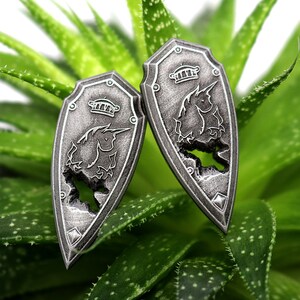May include: Two silver-colored shield-shaped earrings with a unicorn design. The unicorn is outlined in the shield and has a crown on its head.
